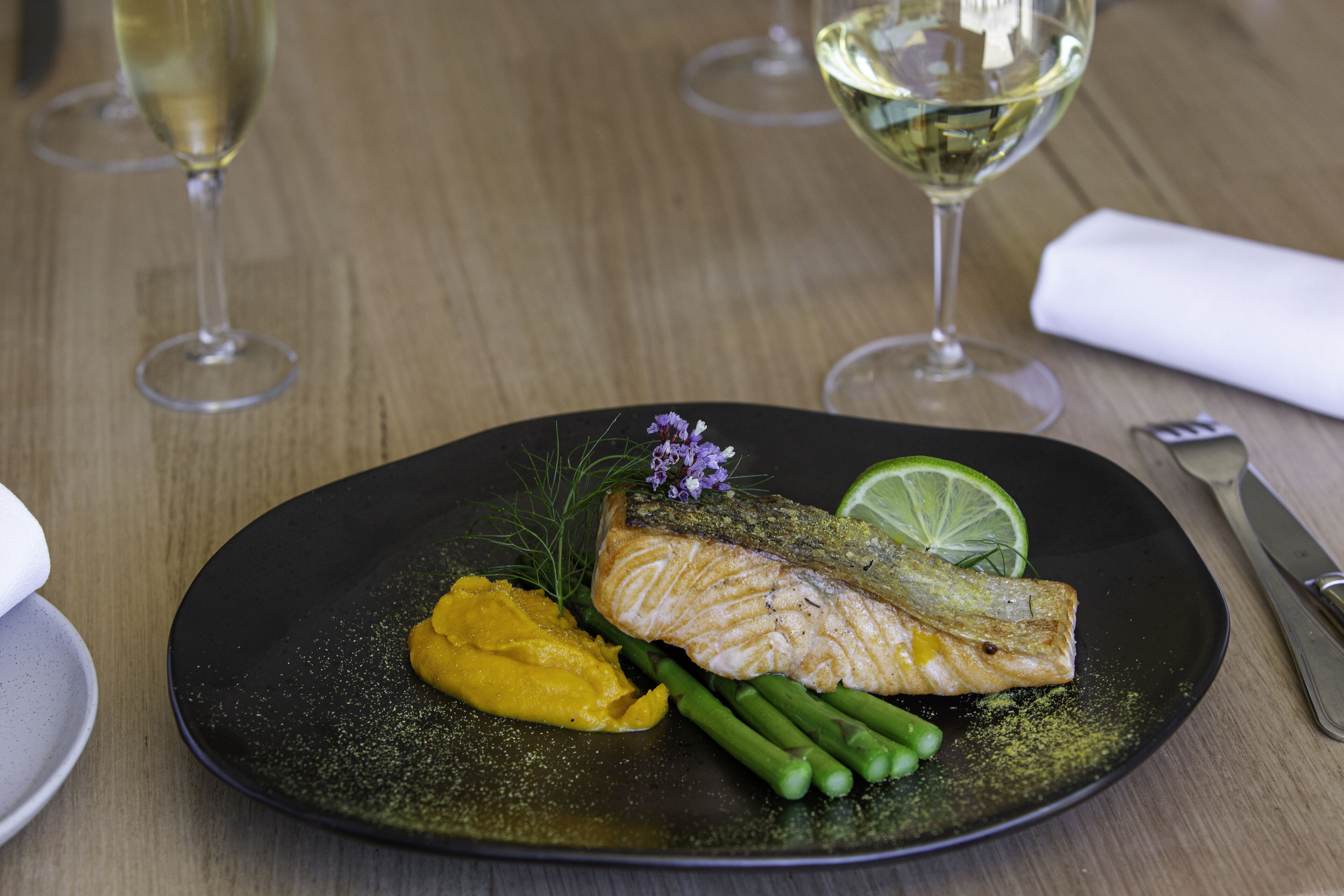 Une assiette noire contenant du saumon, des asperges et un quartier de citron vert.