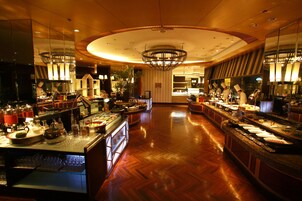 Un vestíbulo de hotel amplio y luminoso con suelo de madera pulida, una araña central y una zona de buffet con diversas estaciones de comida y bebida.