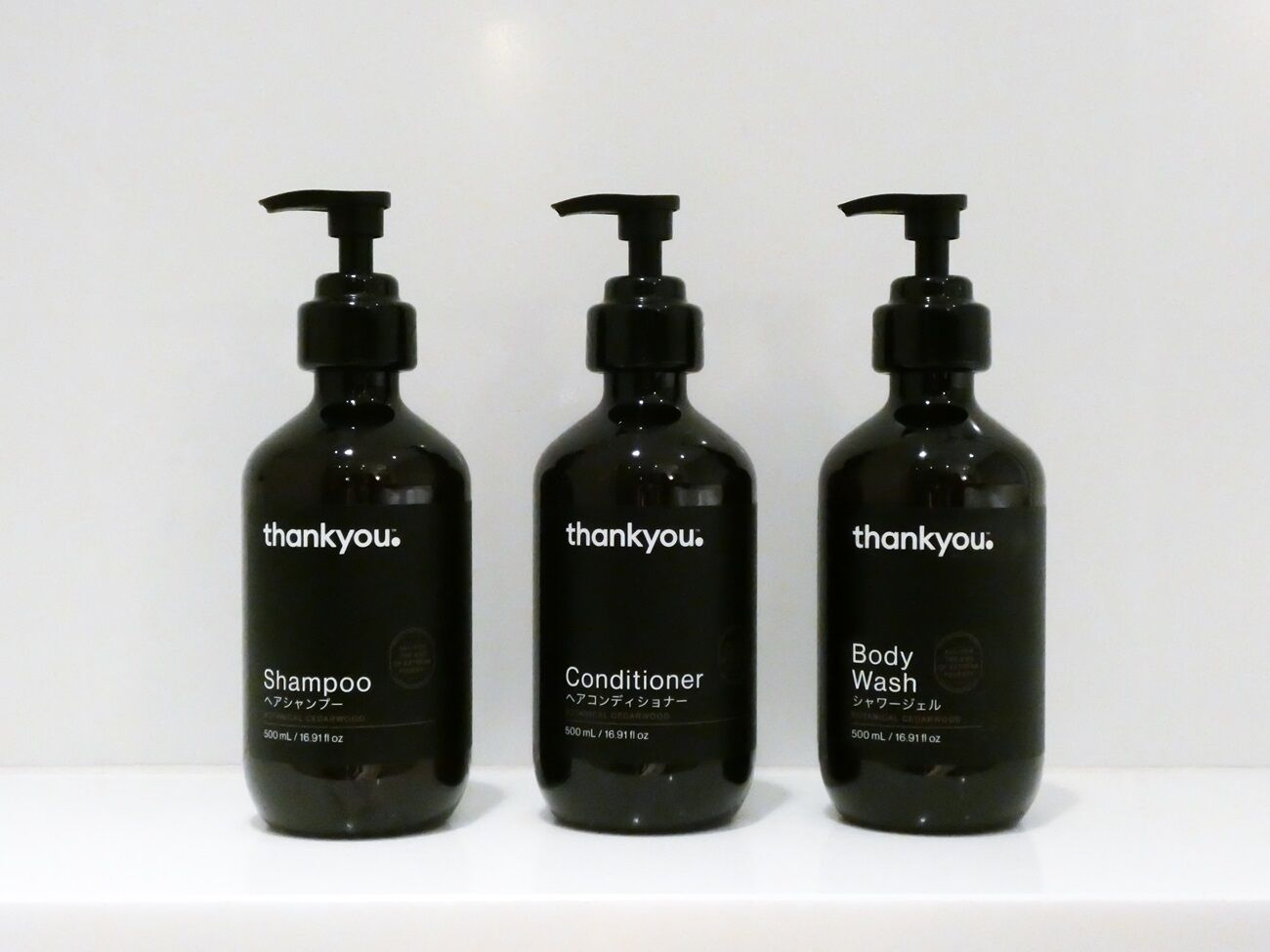Three black bottles of 'thankyou.' shampoo, conditioner, and body wash on a white surface.