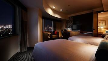 A hotel room with two beds, a desk, a TV, and a view of the city at night.