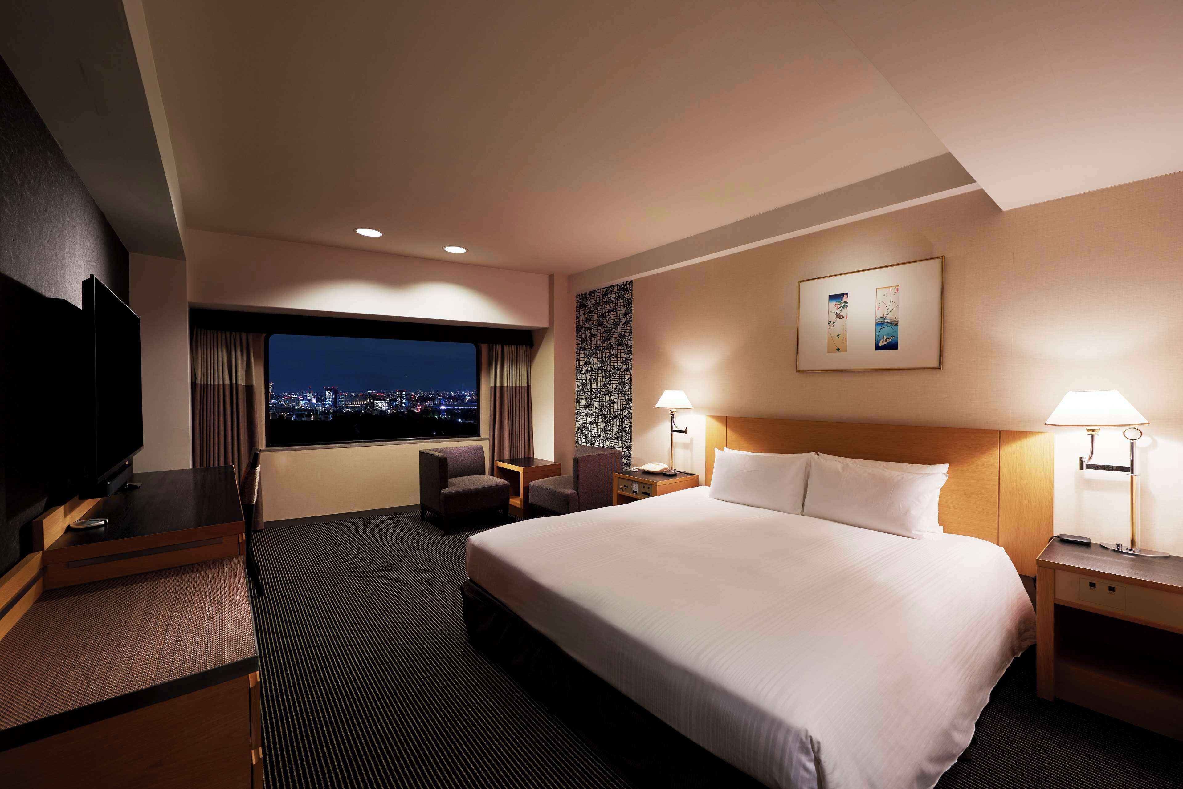 Ein Hotelzimmer mit einem großen Bett, einem Schreibtisch mit Fernseher, zwei Stühlen, einem Nachttisch mit Lampe und einem Fenster mit Blick auf die Stadt.