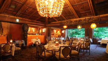 A restaurant with round tables and chairs, a high ceiling with a wooden structure, and a view of greenery outside.