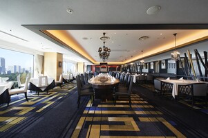 A hotel dining area with a large round table, multiple chairs, and a city view.