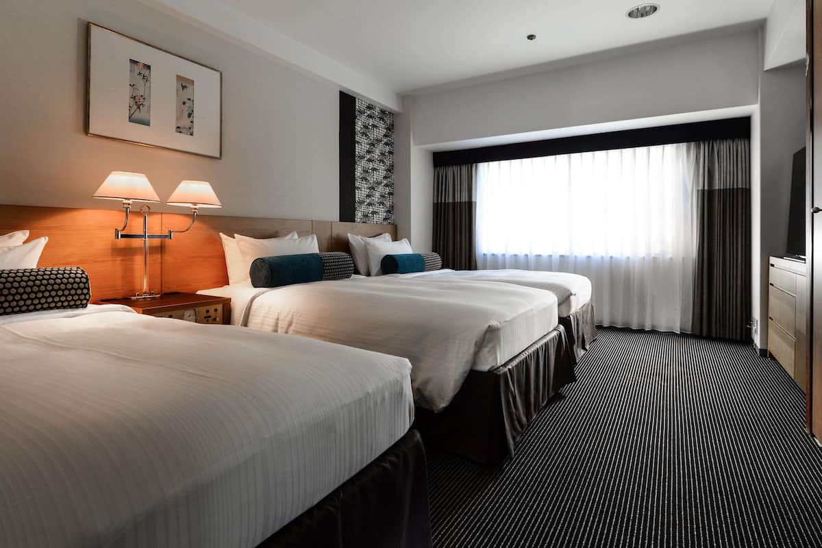 Une chambre d’hôtel avec deux lits, une tête de lit dotée d’un luminaire, une fenêtre avec des rideaux et une œuvre d’art accrochée au mur.