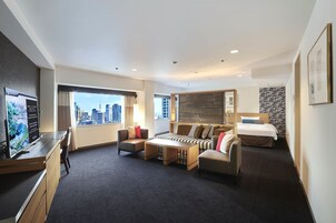 A modern hotel room with a city view, a desk with a computer, a sofa, a bed, and a small table.