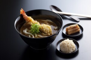 A bowl of ramen with a shrimp, garnished with green onions, and a small bowl of rice on a dark surface.