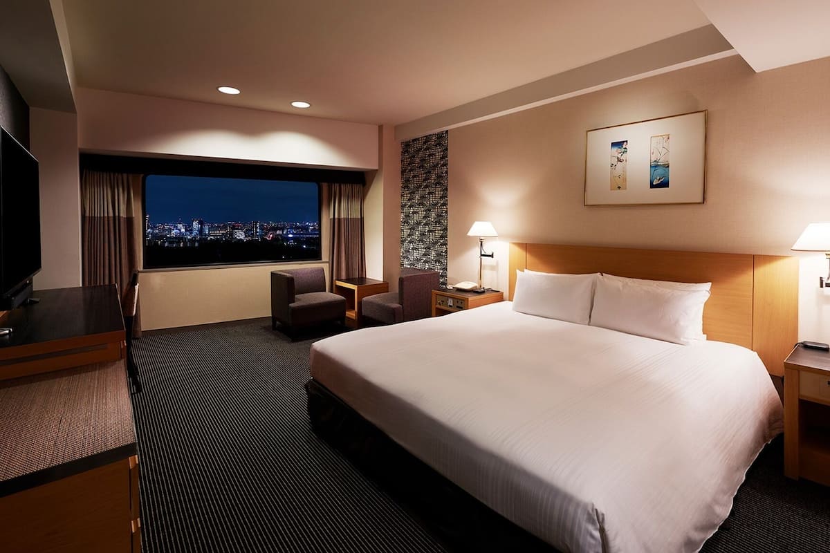 A hotel room with a large bed, a desk with a TV, a chair, a nightstand, and a window with a city view.