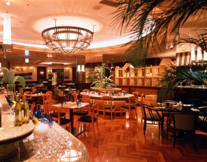 Restaurante com balcão central, várias mesas para refeições e cadeiras. Há plantas e um candelabro à vista.