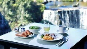 Une table dressée avec des aliments pour le petit-déjeuner, notamment des croissants, une salade et une assiette contenant des œufs et des pommes de terre, le tout disposé devant une vue pittoresque sur une cascade.