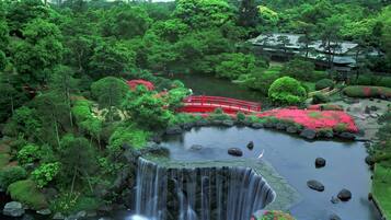 A lush garden with a waterfall, a red bridge, and traditional architecture.