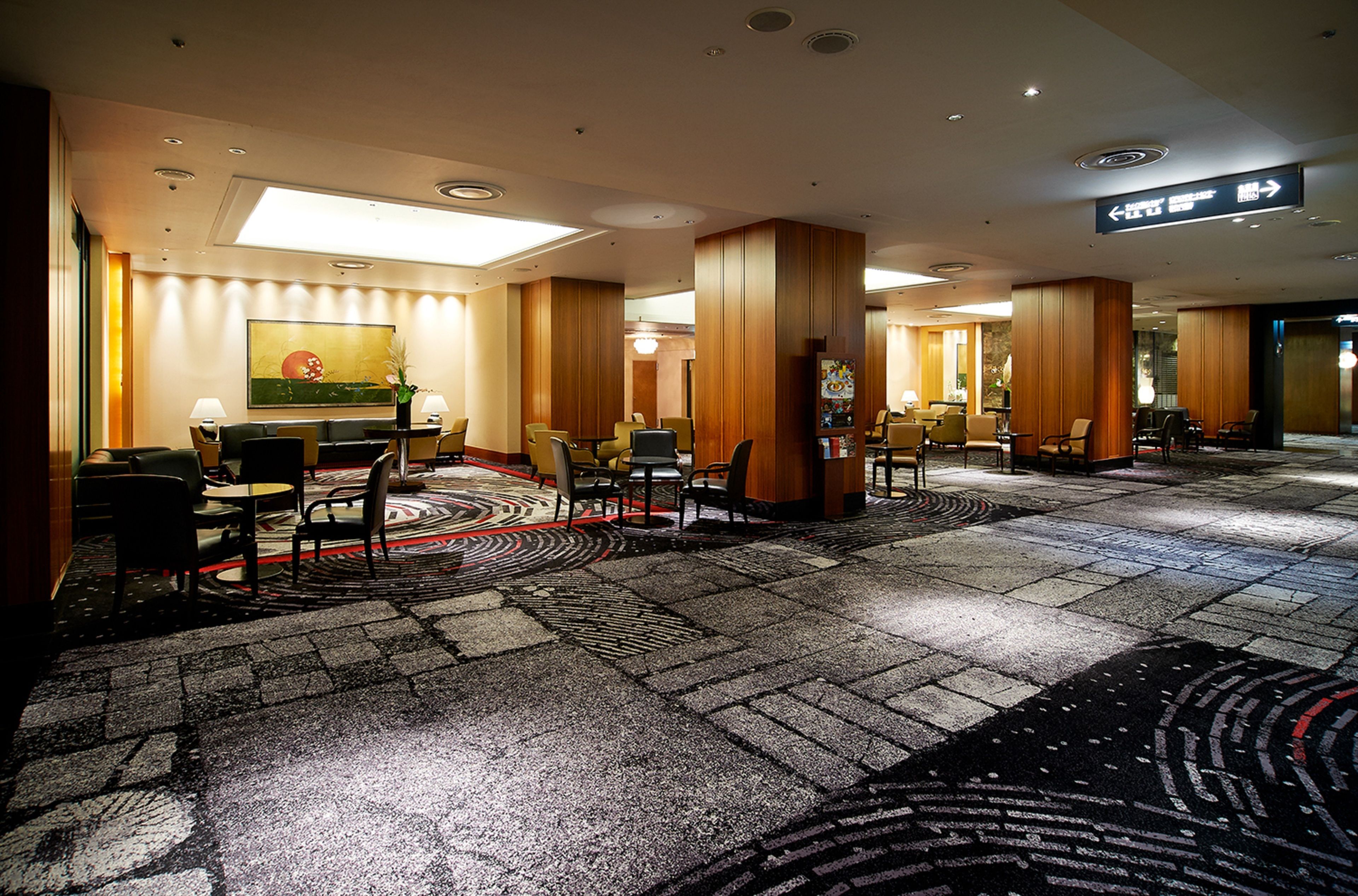 A hotel lobby with a central reception desk, multiple seating areas with chairs and tables, a patterned carpet, and wooden paneling on the walls.