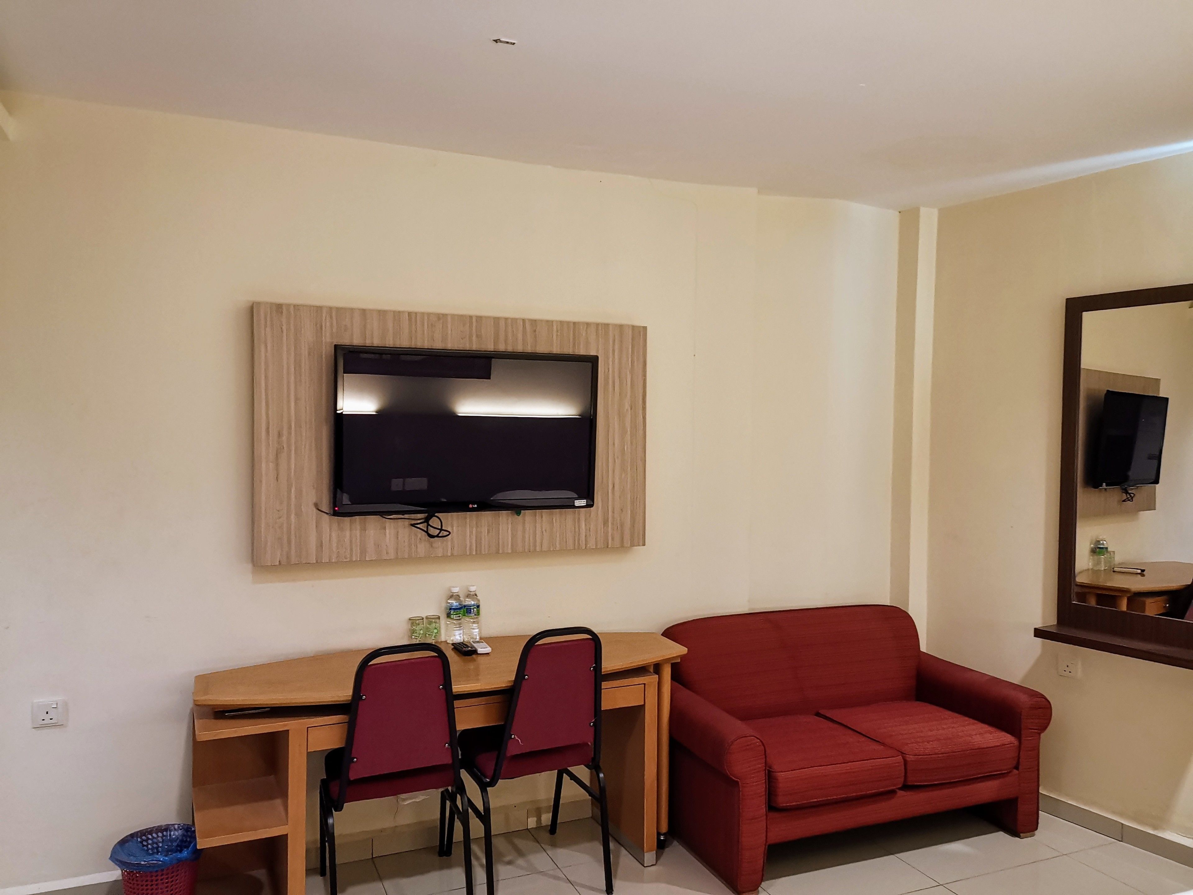 Una habitación con un televisor montado en la pared, un escritorio de madera con dos sillas, un sofá rojo y una mesita con un espejo.
