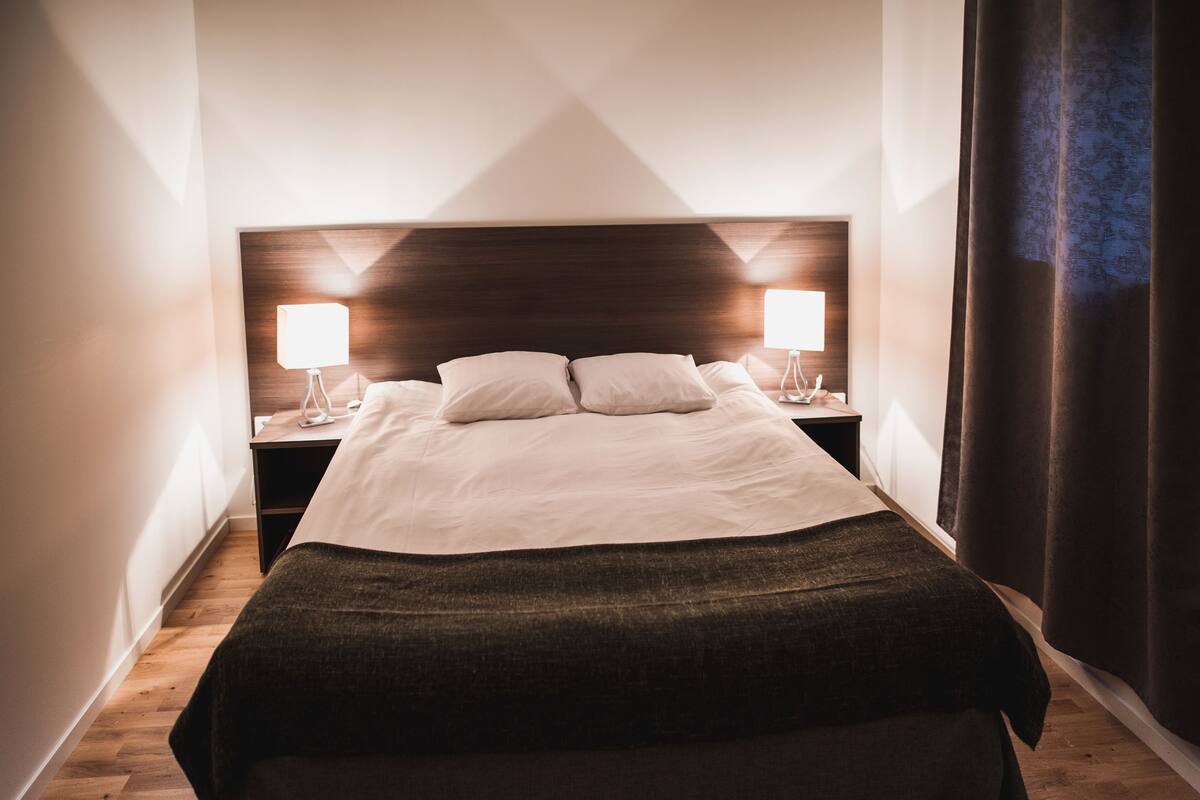 En snyggt bäddad säng med ett mörkt överkast, två sänglampor och en sänggavel i trä.