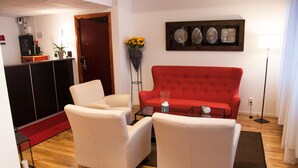 Un soggiorno con un divano rosso, due poltrone bianche, un tavolino da caffè e una lampada da terra. Si apre una porta su un’area cucina con un pensile nero e un tappeto rosso.