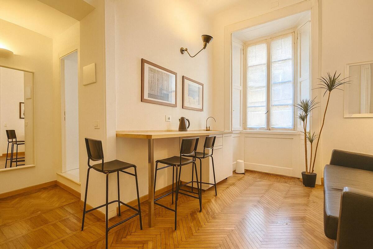 A modern kitchen with a wooden herringbone floor, a dining area with three chairs, a window with blinds, and a potted plant.