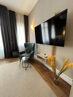 Una sala de estar moderna con un televisor de pantalla plana, un sillón verde azulado, una mesita de centro y un jarrón con plantas secas.