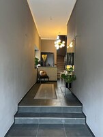 Un pasillo estrecho con piso oscuro, una pequeña zona de estar con una silla y un banco, un radiador y un espejo en la pared.