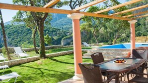 Un espacio al aire libre techado con mesa y sillas, piscina y vista a un paisaje montañoso.