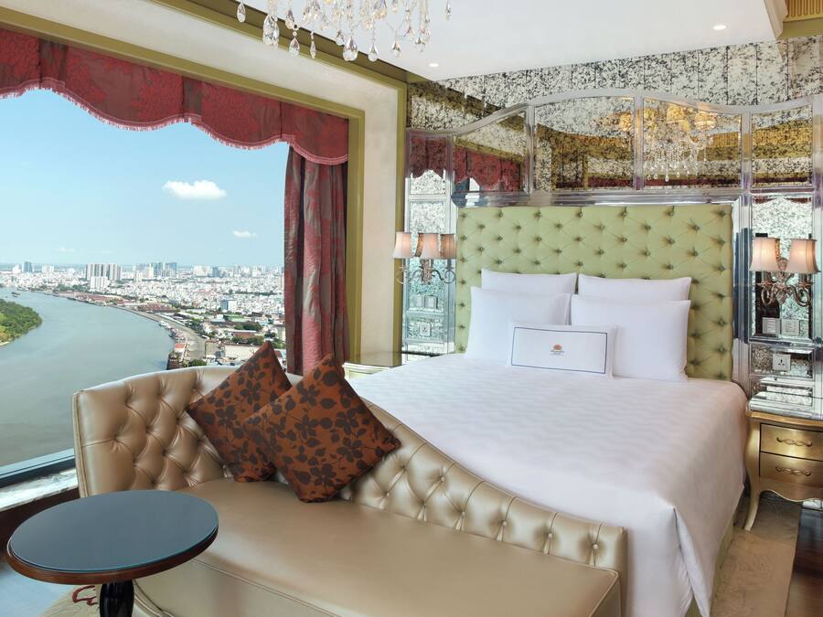 A luxurious bedroom with a large bed, a chaise lounge, a chandelier, and a view of a cityscape.