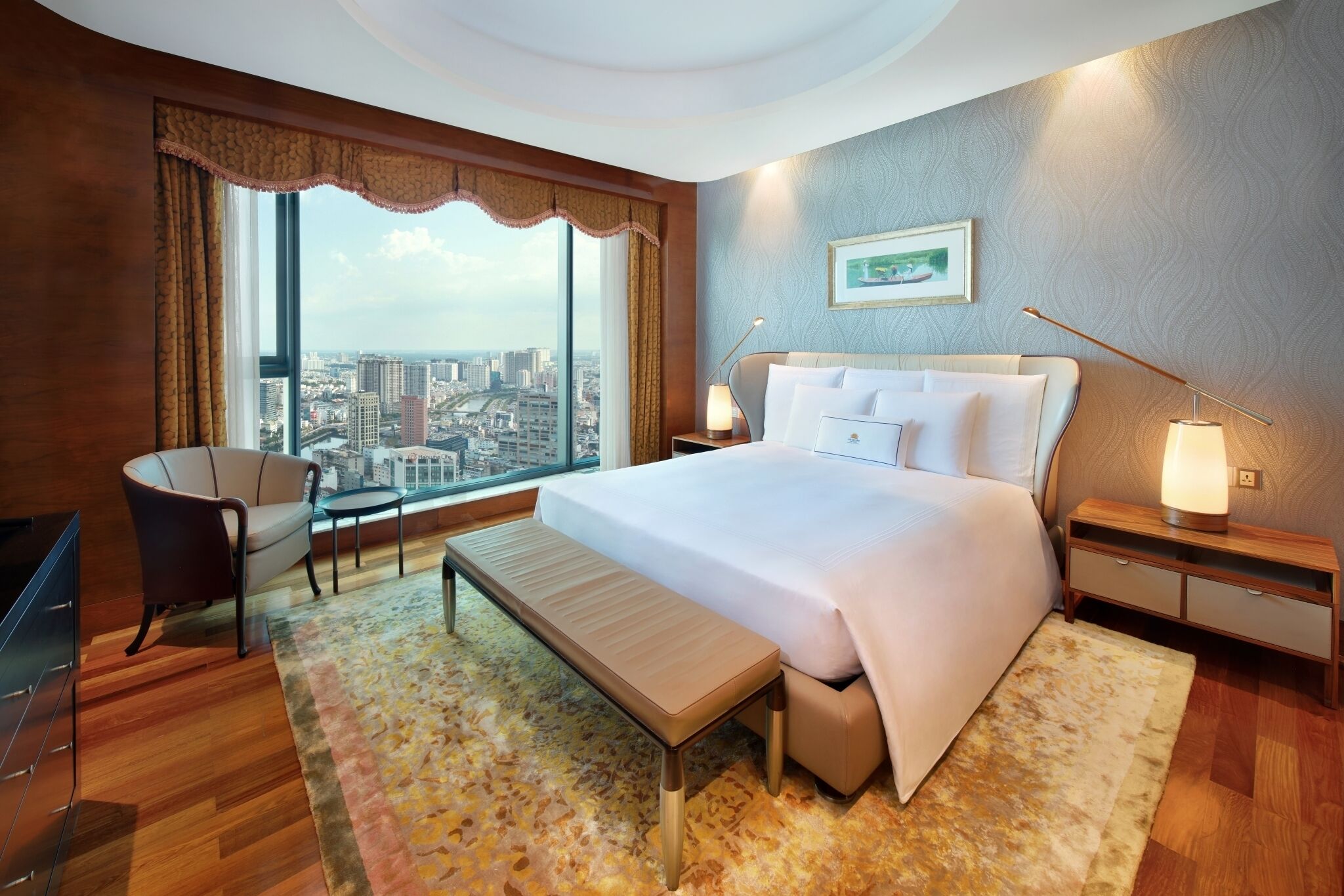 A modern hotel room with a large bed, a sitting area with a chair, a bench, and a view of the city through large windows.