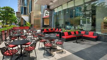An outdoor seating area with tables and chairs, red cushioned sofas, and a large glass wall reflecting the surroundings.