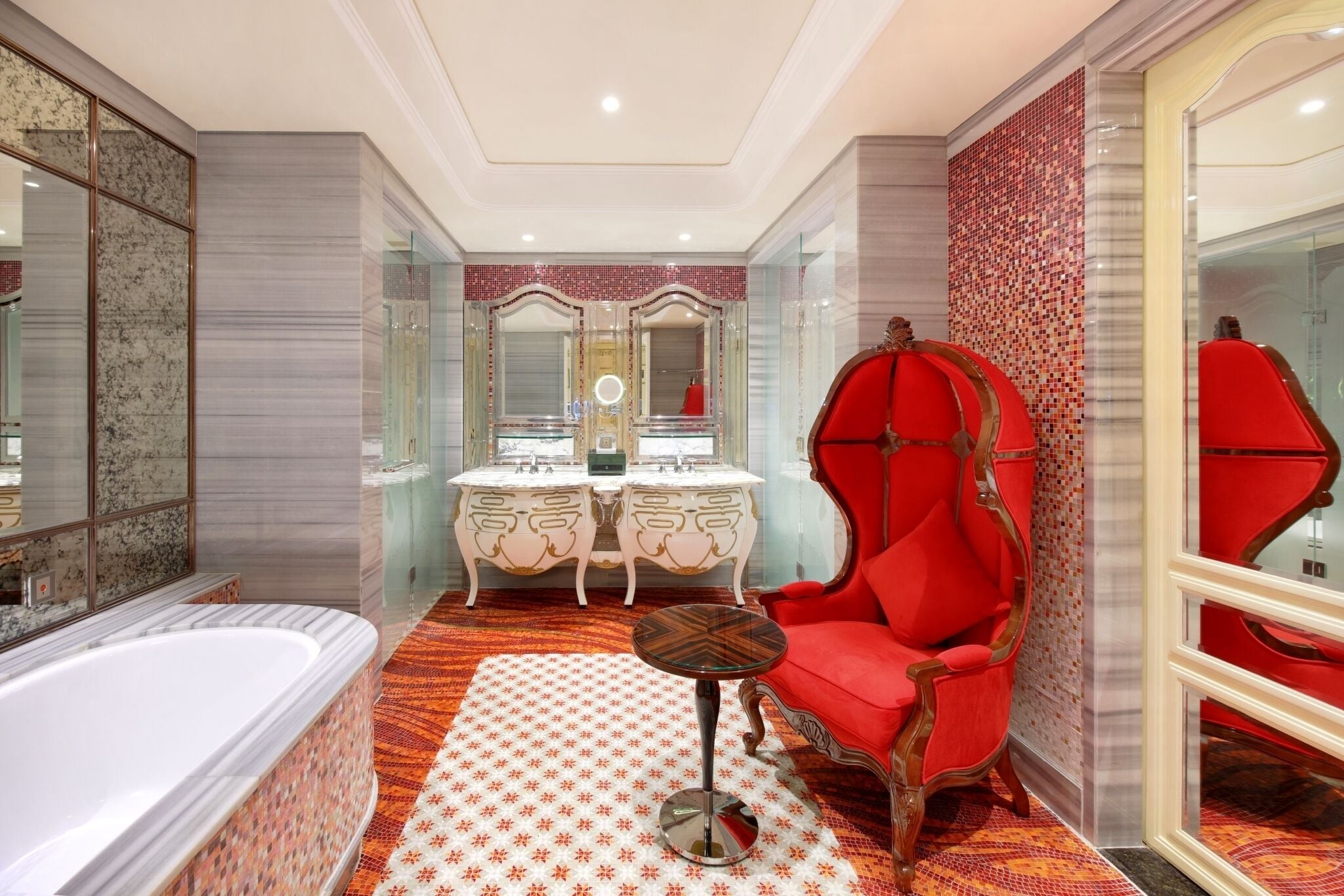 A luxurious bathroom with a large bathtub, two ornate vanity tables, a red upholstered chair, and a patterned rug.
