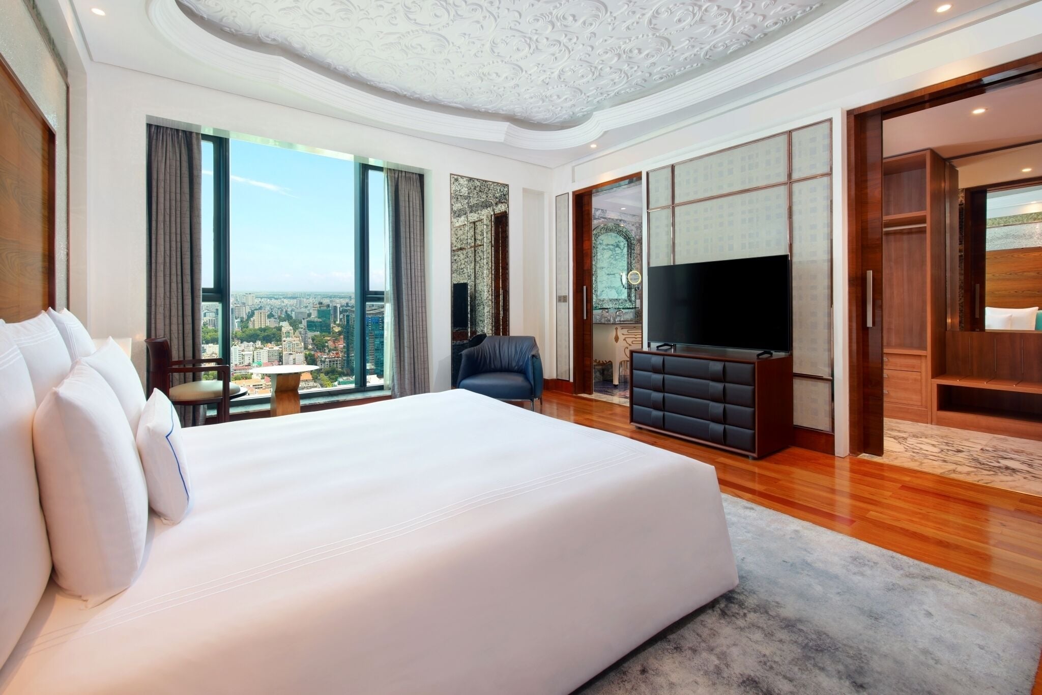 A spacious hotel room with a large bed, a flat-screen TV, a seating area with a table, and a view of the city.