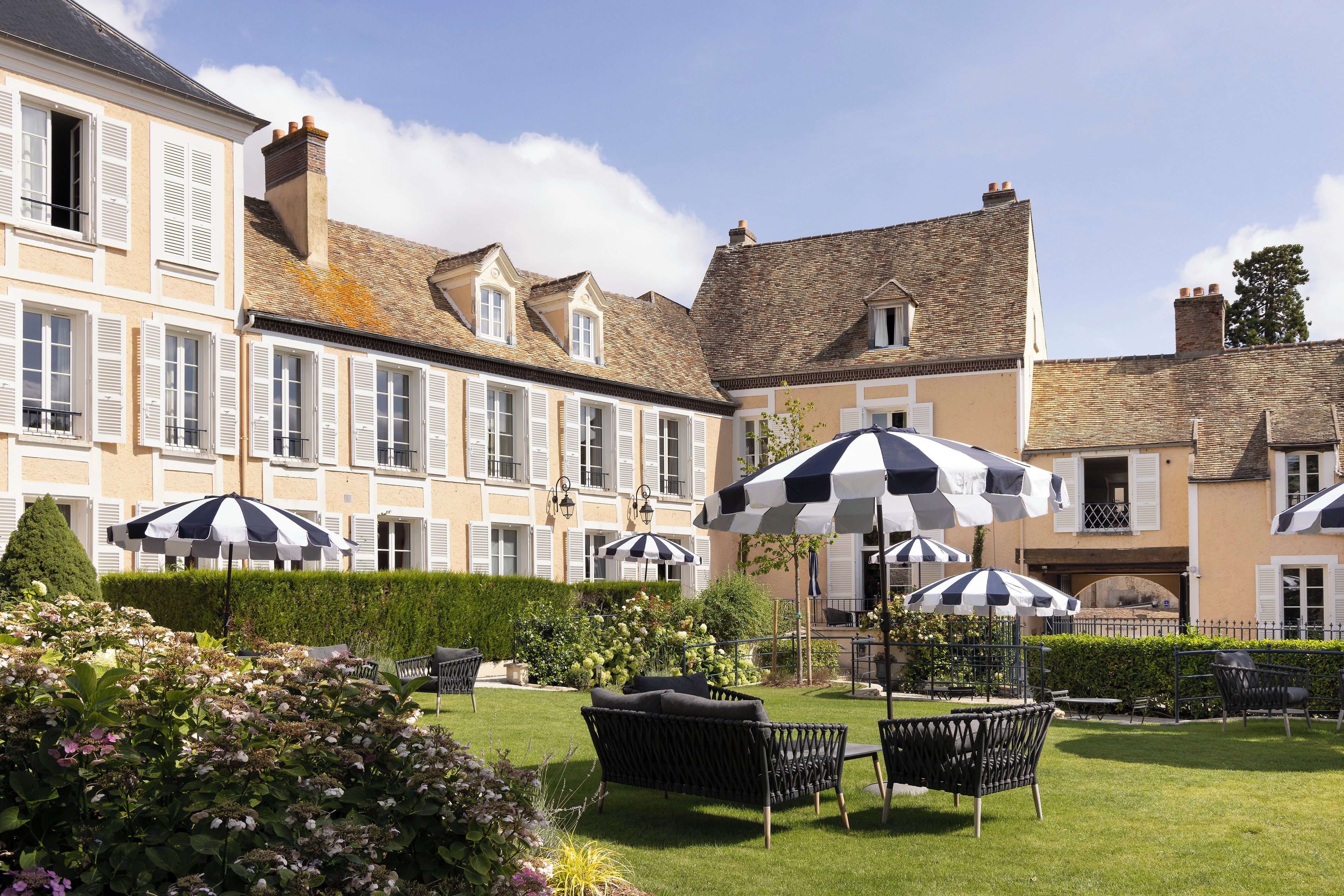 A courtyard with outdoor seating, a hedge, and a building with multiple windows and shutters.