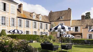 A courtyard with outdoor seating, a hedge, and a building with multiple windows and shutters.