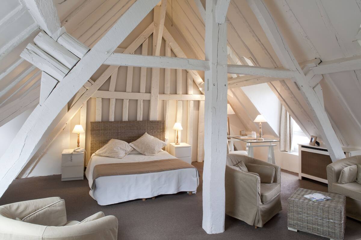 Een gezellig slaapkamer onder de dakrand met een groot bed, twee nachtkastjes met lampen, een rieten stoel, een bank en een kleine tafel.
