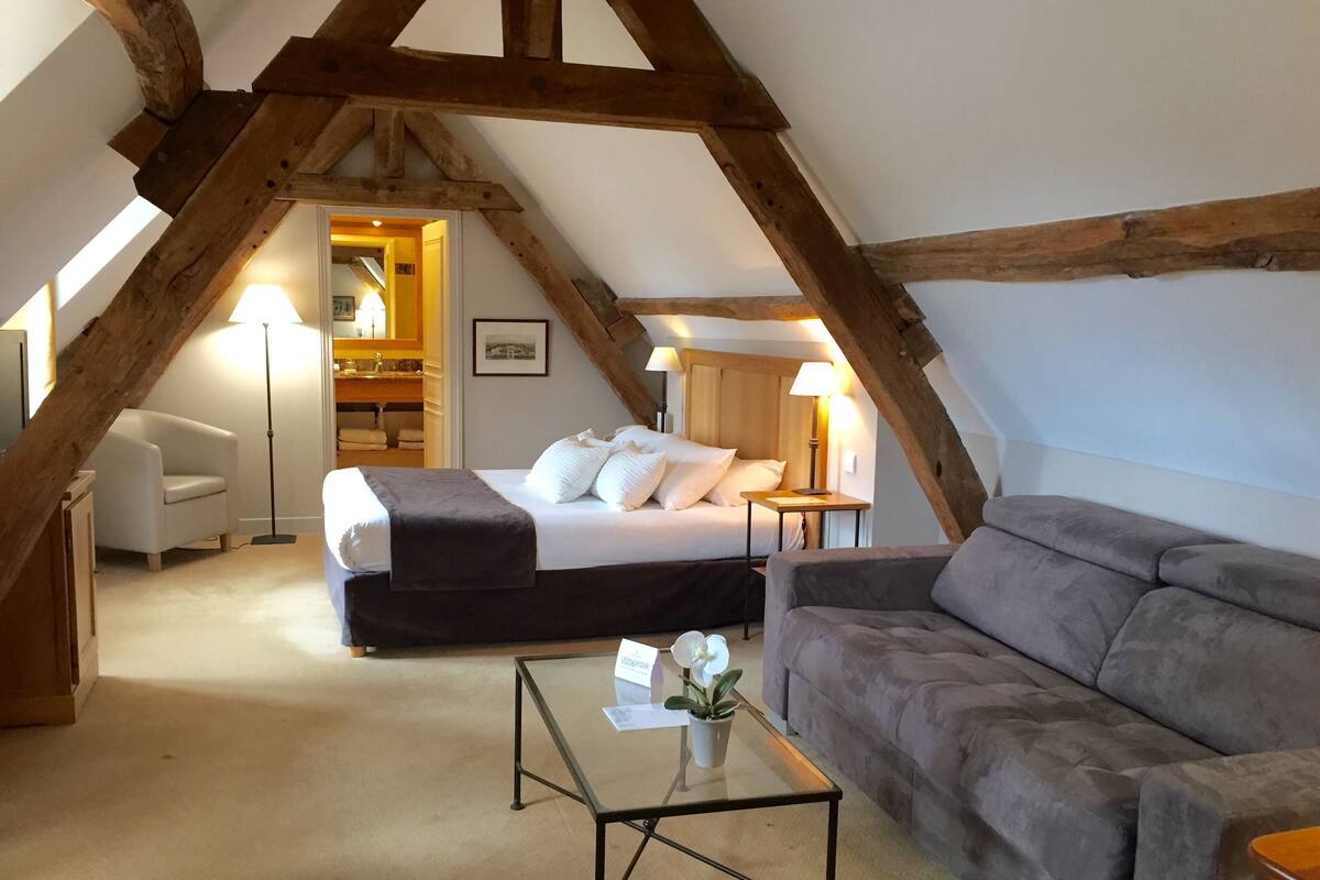Een gezellige kamer met een bed, een grijze bank, een glazen bijzettafeltje en een houten plafondconstructie.