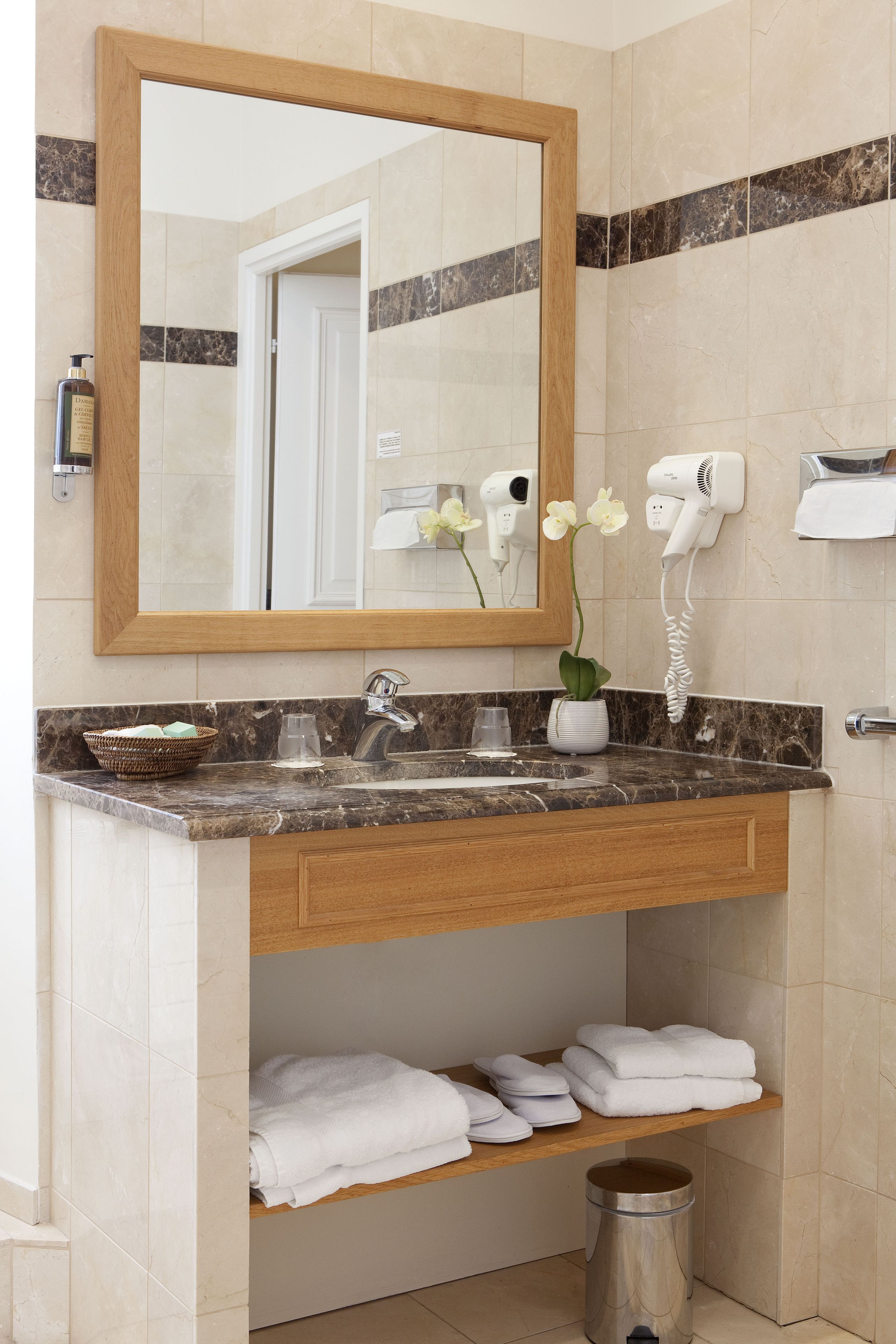 Une salle de bain avec un lavabo en marbre, un meuble-lavabo en bois, un grand miroir, un sèche-cheveux et une poubelle.