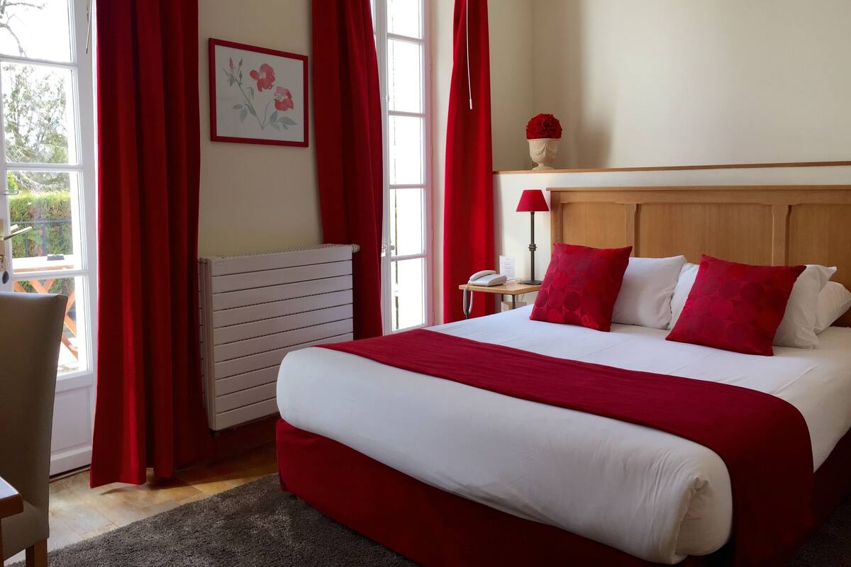 Een hotelkamer met een groot bed, rode gordijnen, een houten hoofdeinde, een nachtkastje met een lamp en een radiator.