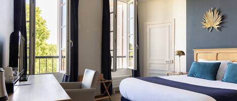 A hotel room with a bed, a desk with a chair, a bedside table, a lamp, and a view of the outdoors.