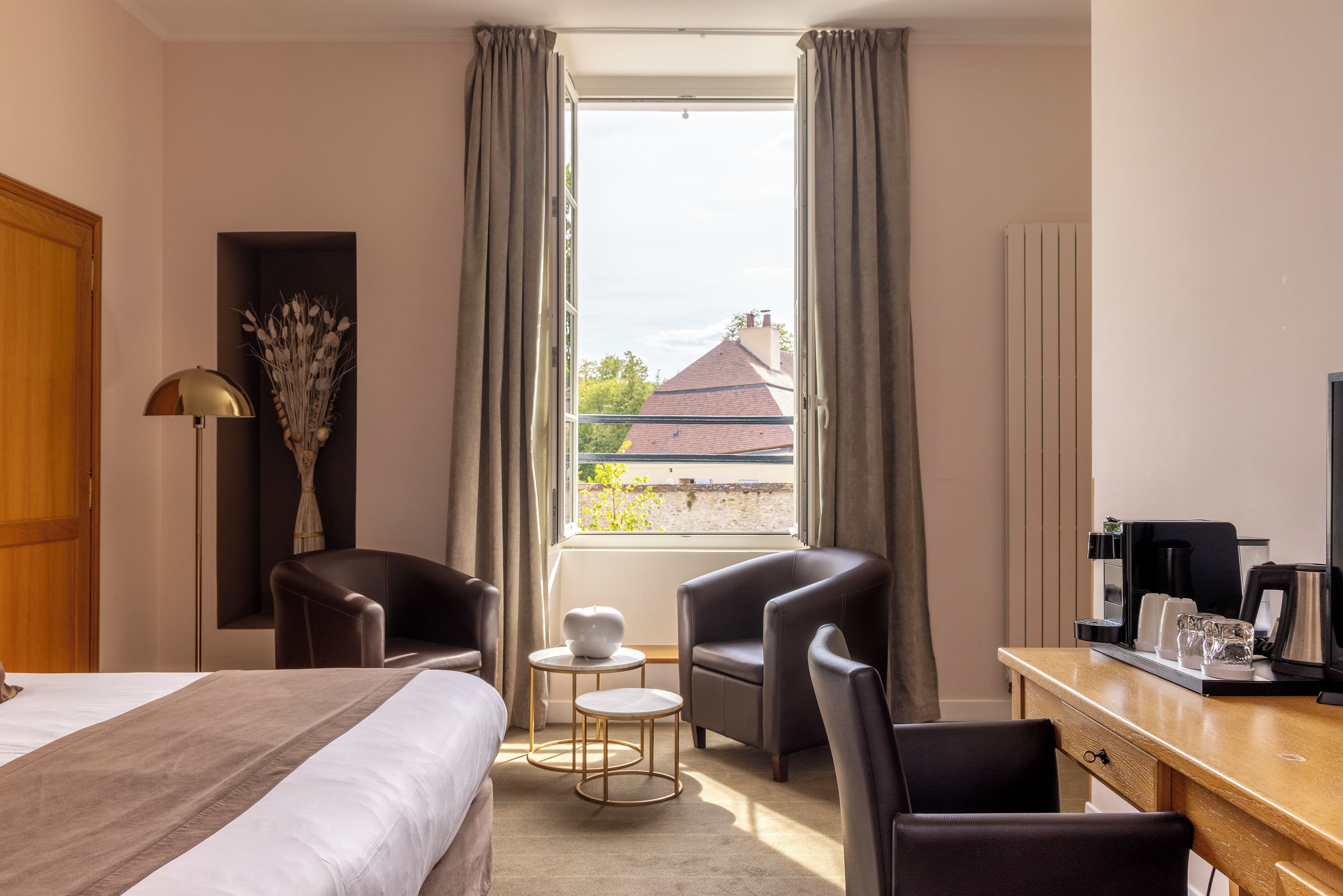A hotel room with a bed, two armchairs, a small table, a coffee station, and a view of buildings outside.