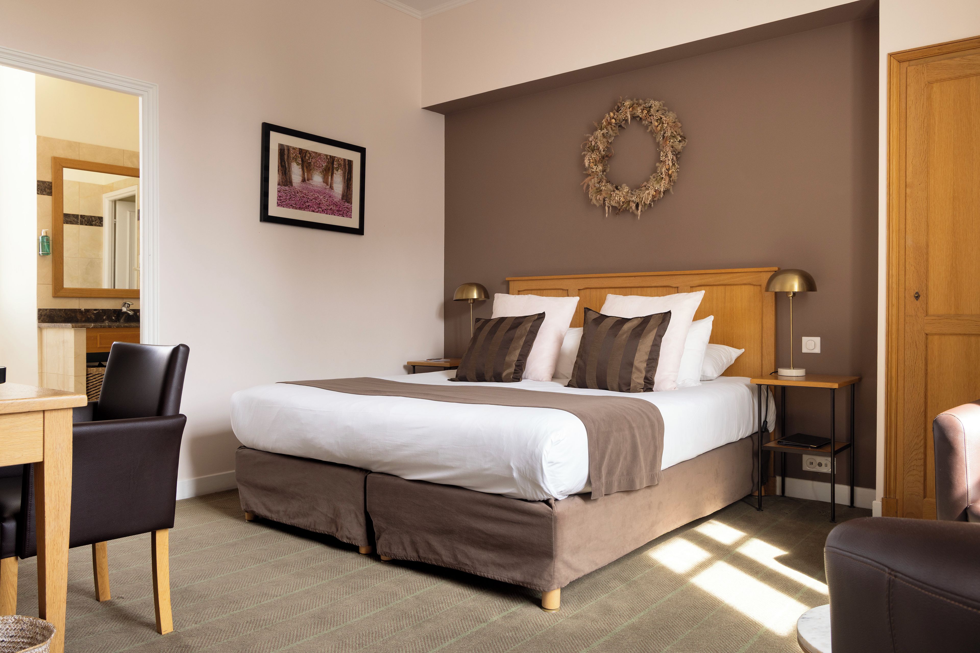 A hotel room with a bed, a desk, a chair, a bedside table, a lamp, and a picture on the wall.