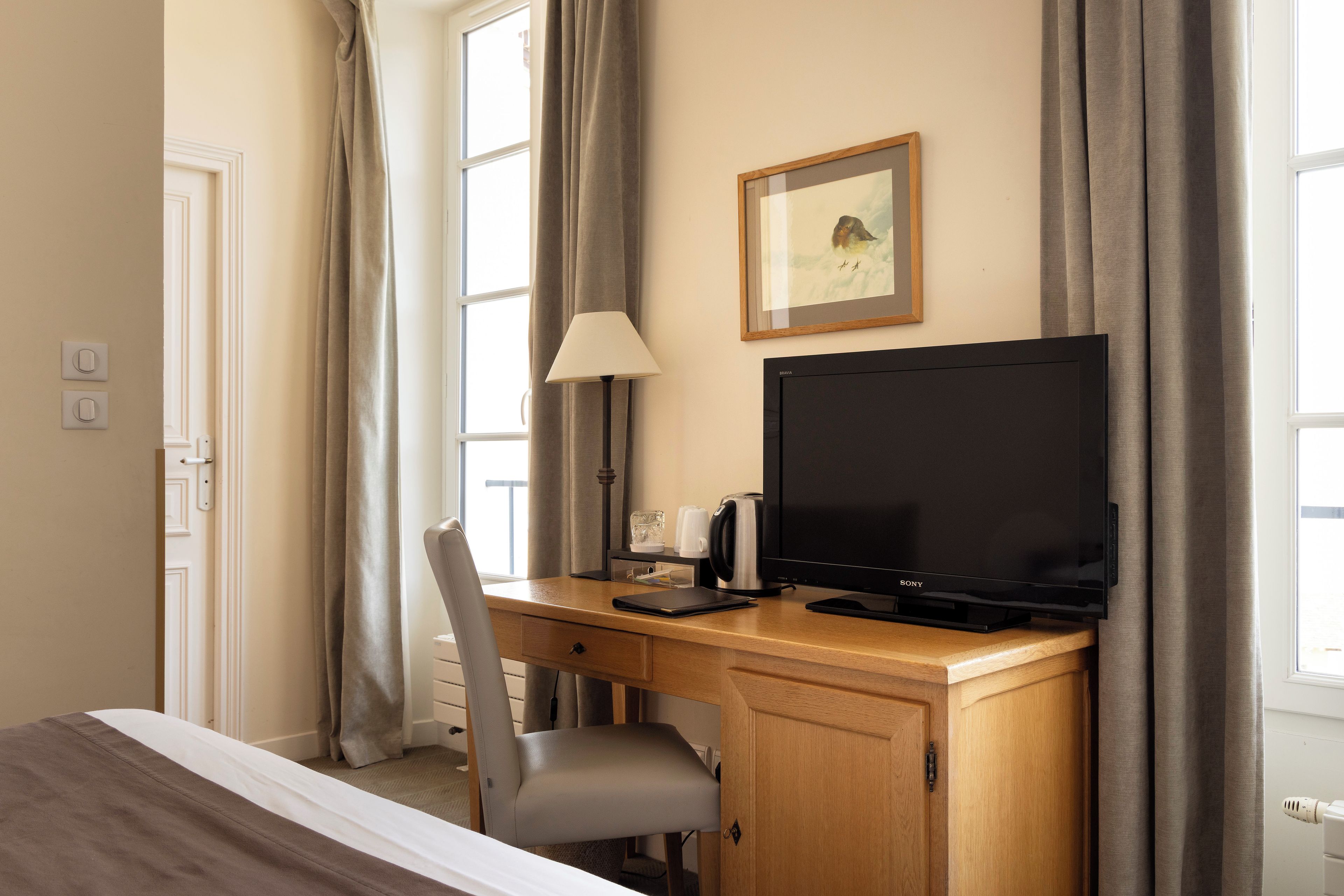 Quarto de hotel com escrivaninha de madeira, televisão preta, luminária e quadro na parede.