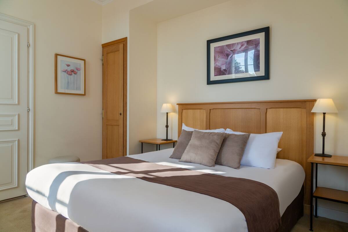 Een netjes opgemaakt bed met een houten hoofdbord, twee nachtkastjes met lampen, en een ingelijst beeld aan de muur.