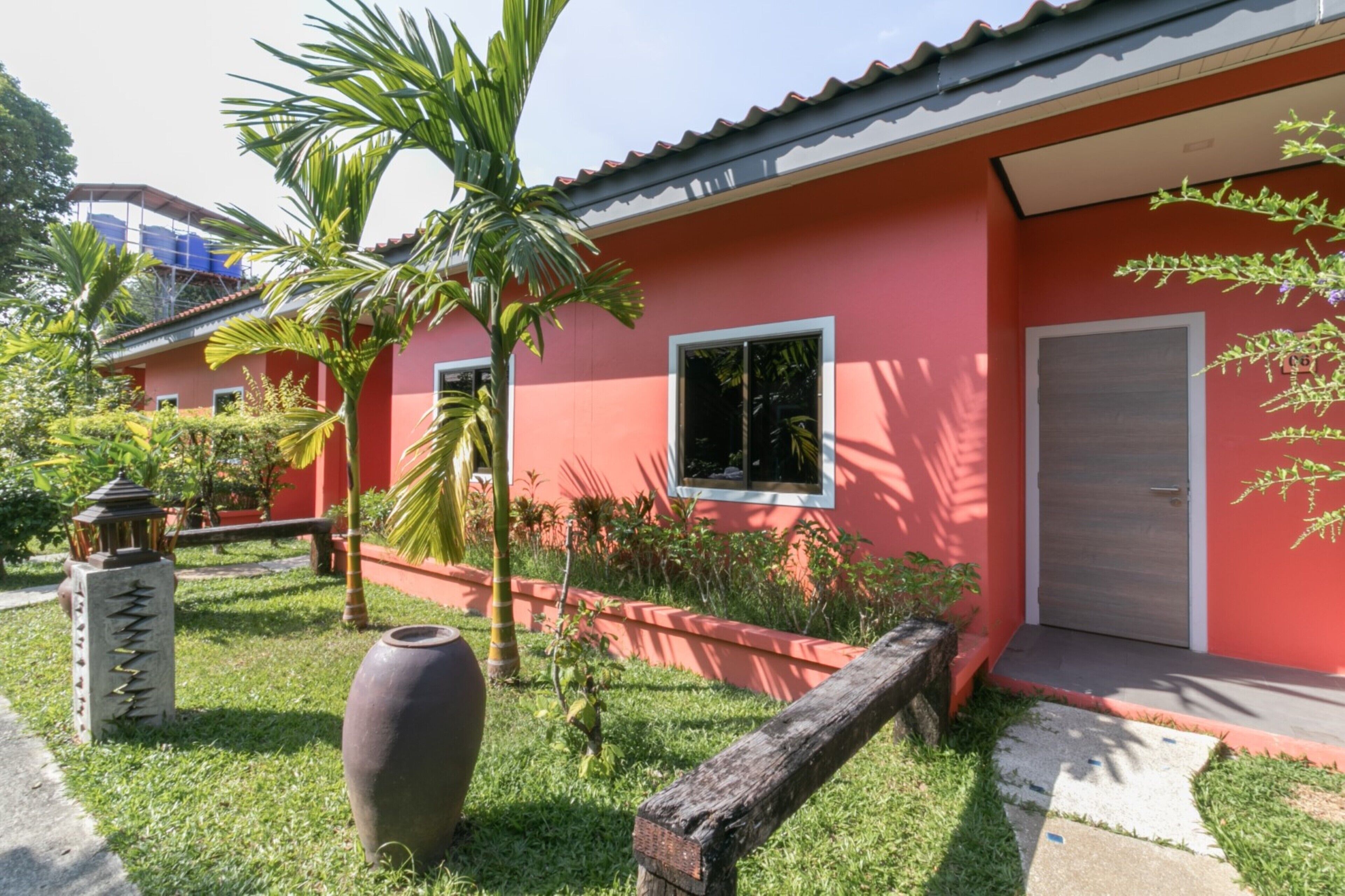 Un bâtiment rouge avec un toit en tuiles, un petit jardin planté de palmiers et un grand vase décoratif.