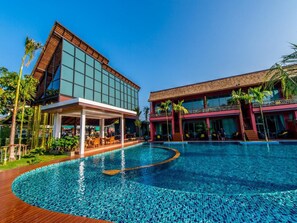 Un hotel moderno con una piscina grande, zona de estar exterior cubierta y exuberante vegetación tropical.