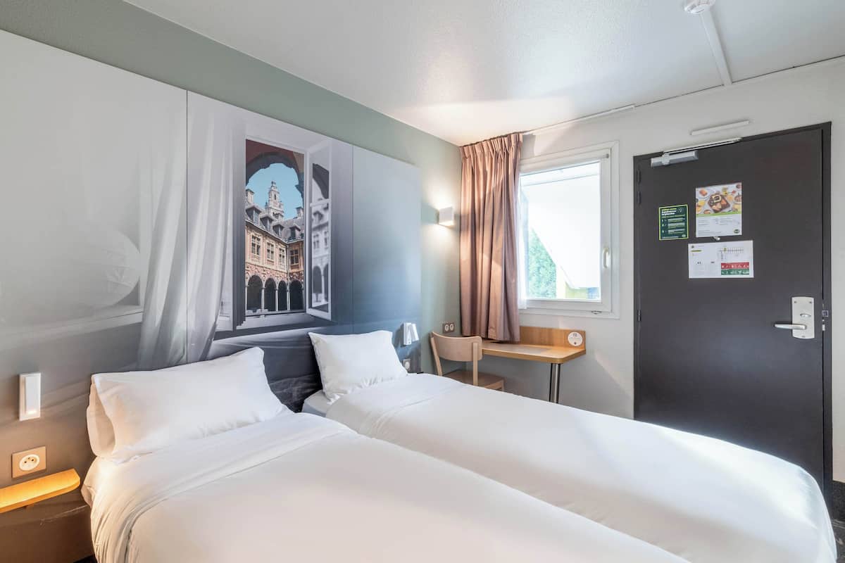 A hotel room with two beds, a desk, a chair, a window with curtains, and a door with a bulletin board.