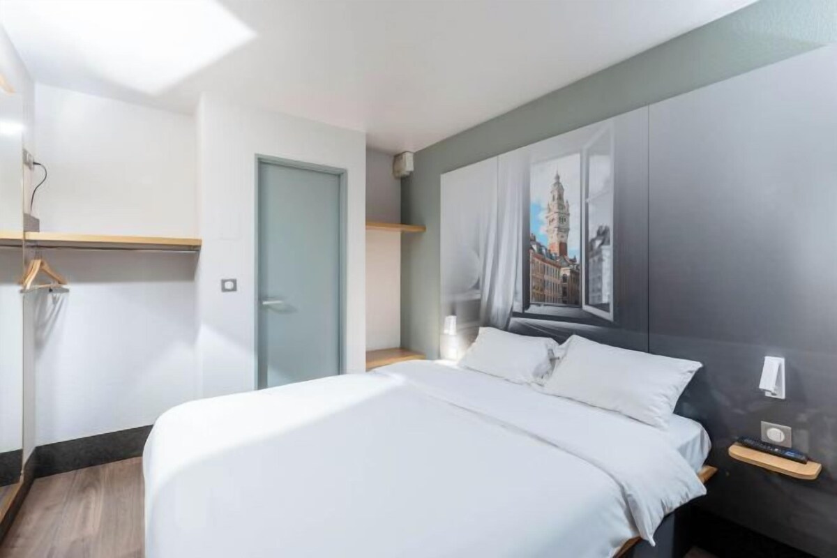 Una habitación de hotel moderna con una cama grande, una mesita de noche, un televisor montado en la pared y un ventanal con vistas a la ciudad.