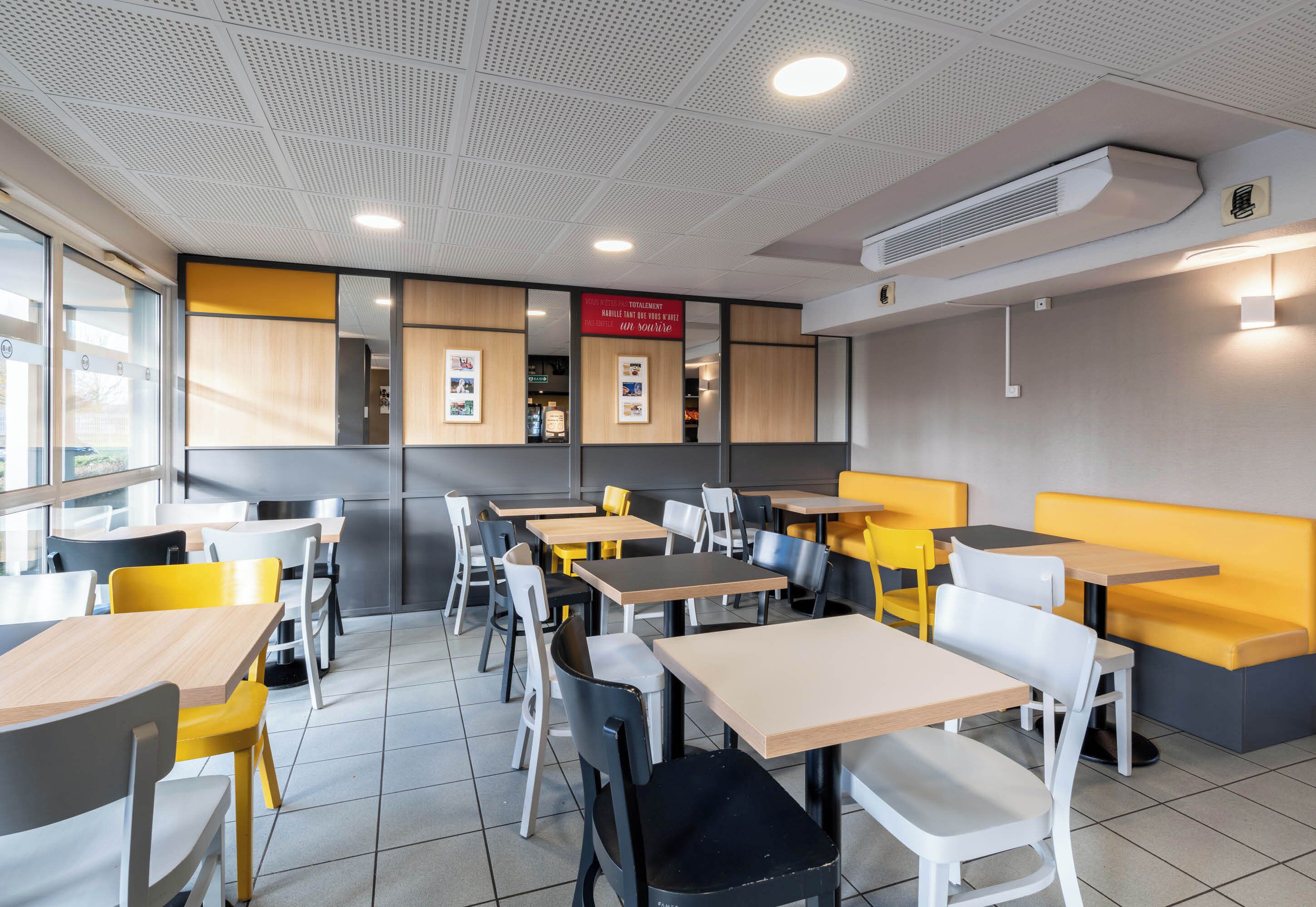 En modern restauranginteriör med flera bord och stolar, ett tak med infälld belysning och en synlig luftkonditioneringsenhet.