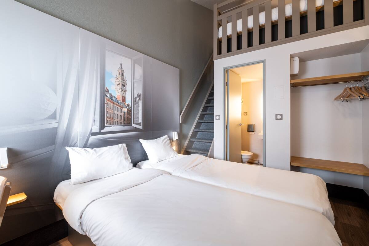 A modern hotel room with a large bed, a staircase, a wardrobe, and a framed cityscape picture.