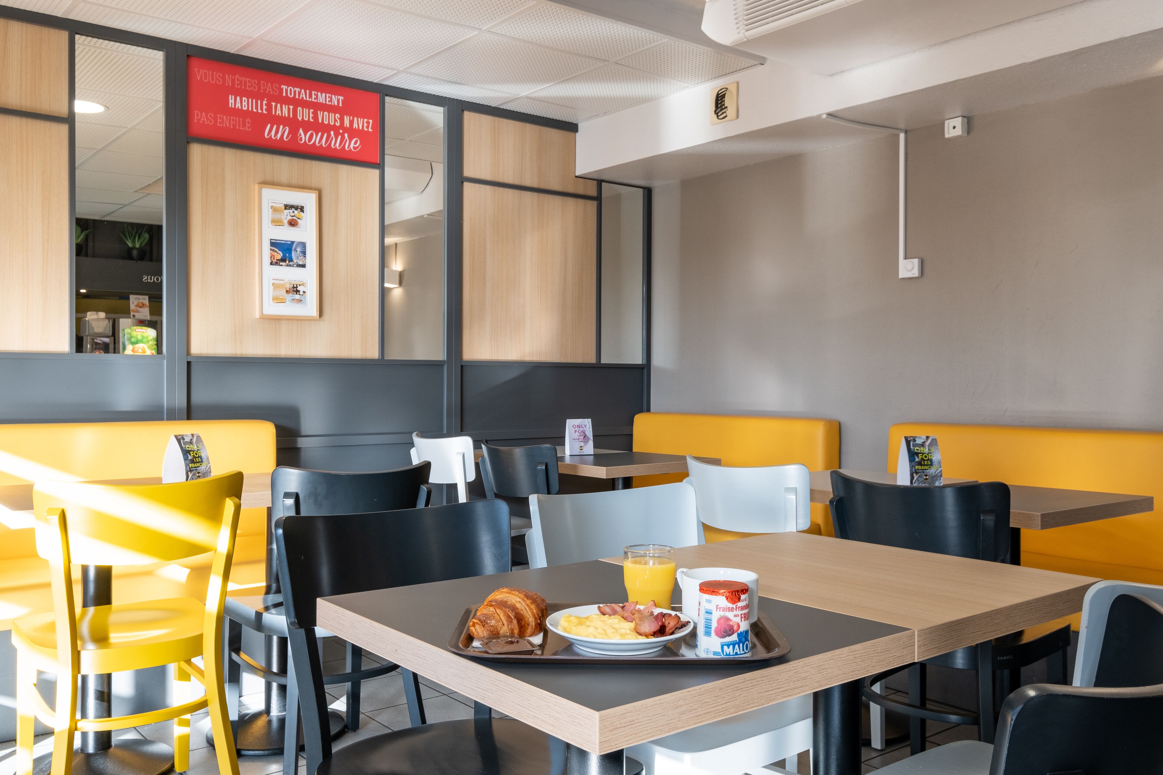 Un coin repas avec des chaises jaunes et noires, des tables en bois et un plateau contenant des croissants, des œufs et du jus d’orange.