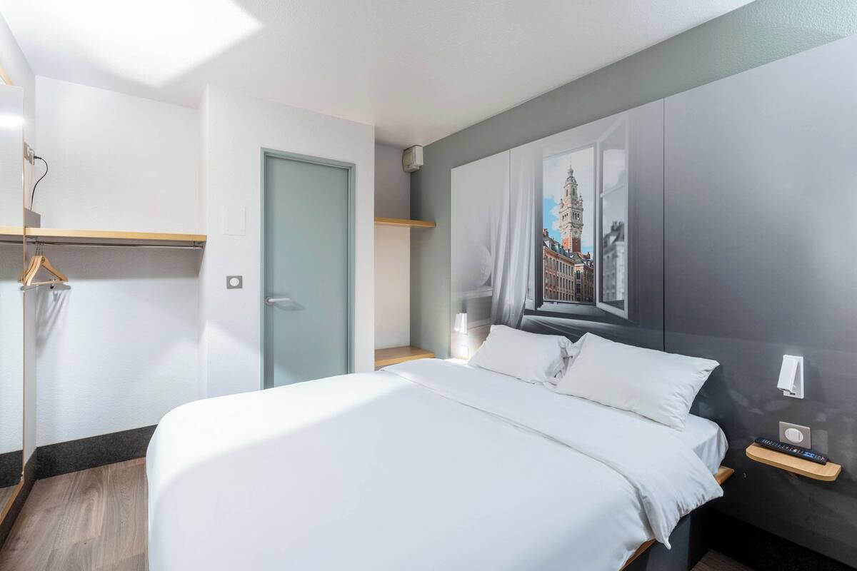 A modern hotel room with a large bed, a wardrobe, a desk with a lamp, and a wall-mounted TV.