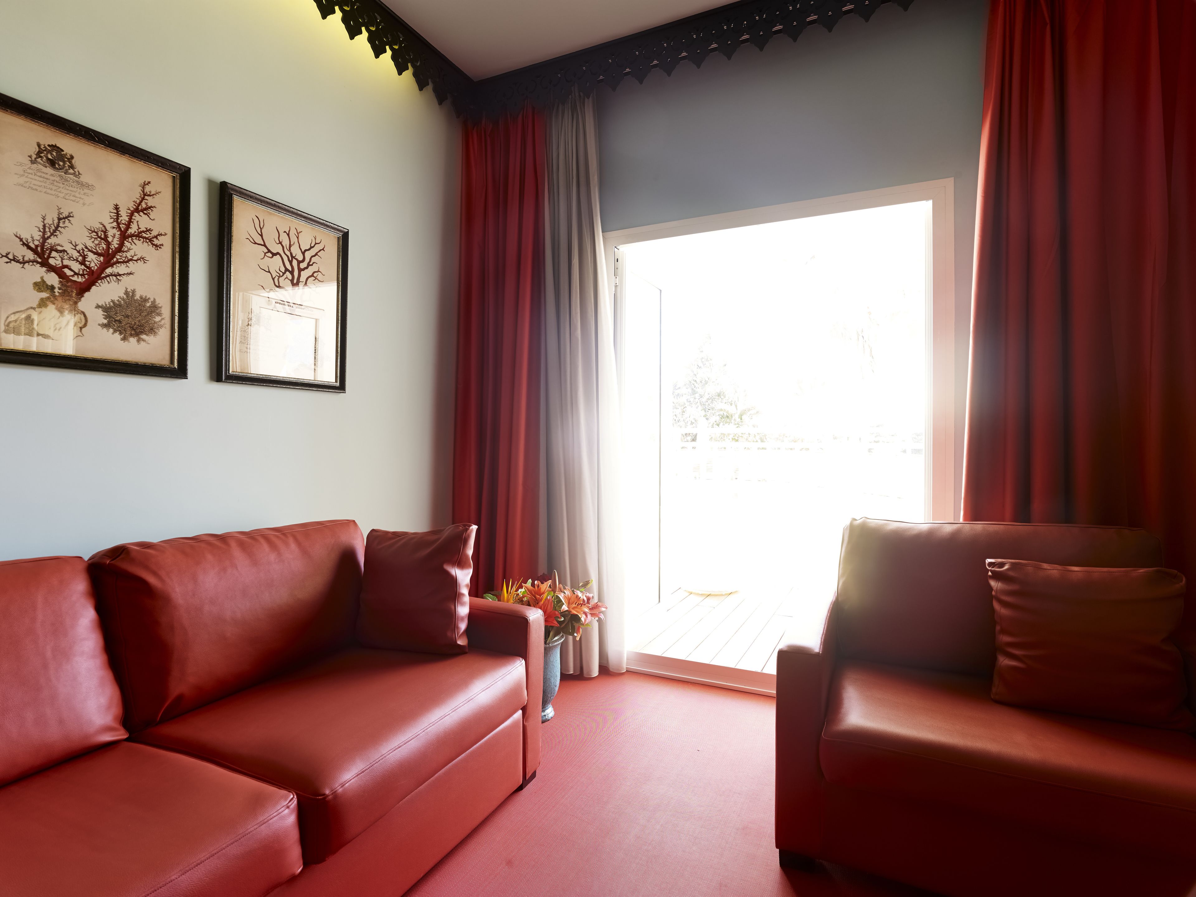 A room with a red leather sofa, a smaller armchair, and a large window with red curtains. There are two framed pictures on the wall.