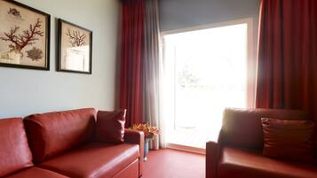 A room with a red leather sofa, a smaller armchair, and a large window with red curtains. There are two framed pictures on the wall.