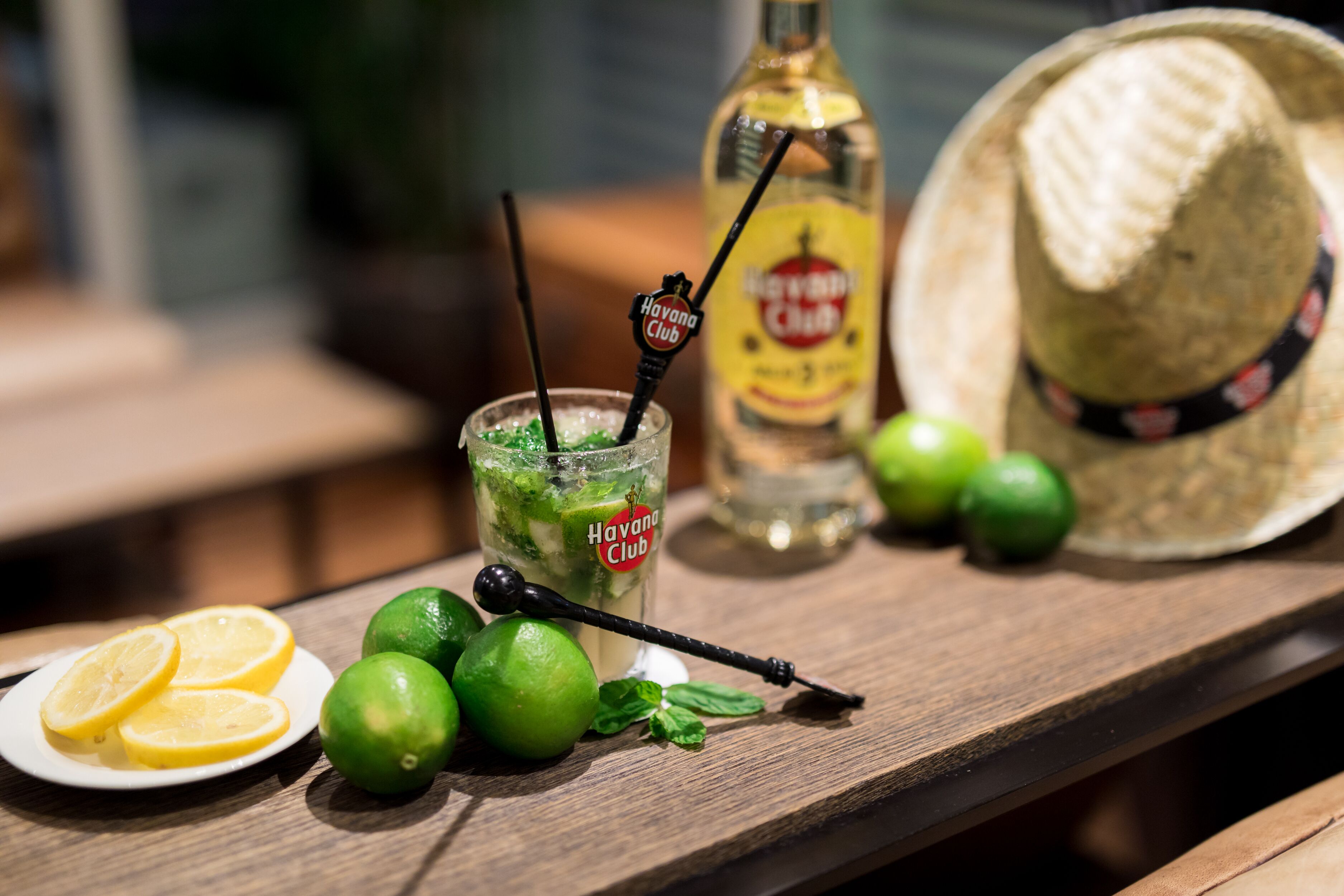 A glass of Havana Club rum cocktail with lime wedges and mint leaves, a bottle of the same rum, and a straw with a Havana Club logo.