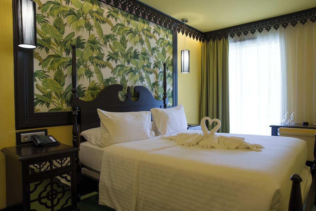 A hotel room with a bed, a nightstand, a wall with a tropical wallpaper, and a window with curtains.
